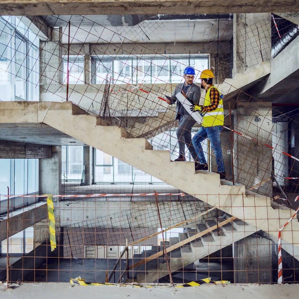 Zwei Bauingenieure stehen auf einer Rohbautreppe und begutachten Baupläne
